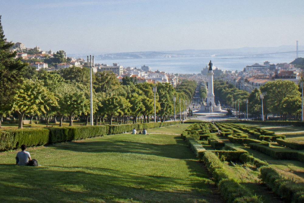 A long grassy park axis with clipped hedges leads downhill toward a monument, avenue trees, and the Tagus beyond.