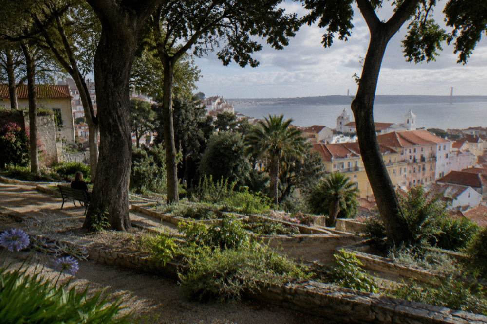 Terraced garden beds with trees overlook Lisbon rooftops and the Tagus estuary.