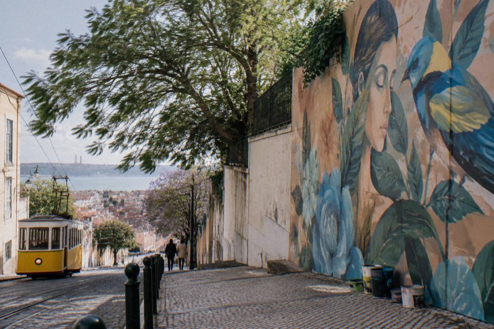 A yellow tram descends a cobbled Lisbon street beside a large mural wall and distant waterfront skyline.