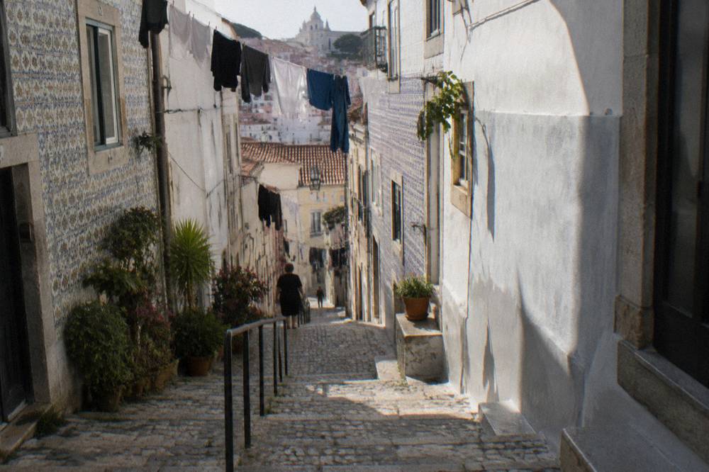 A narrow steep cobbled Lisbon lane with tiled walls, laundry lines overhead, potted plants, and a distant city view.