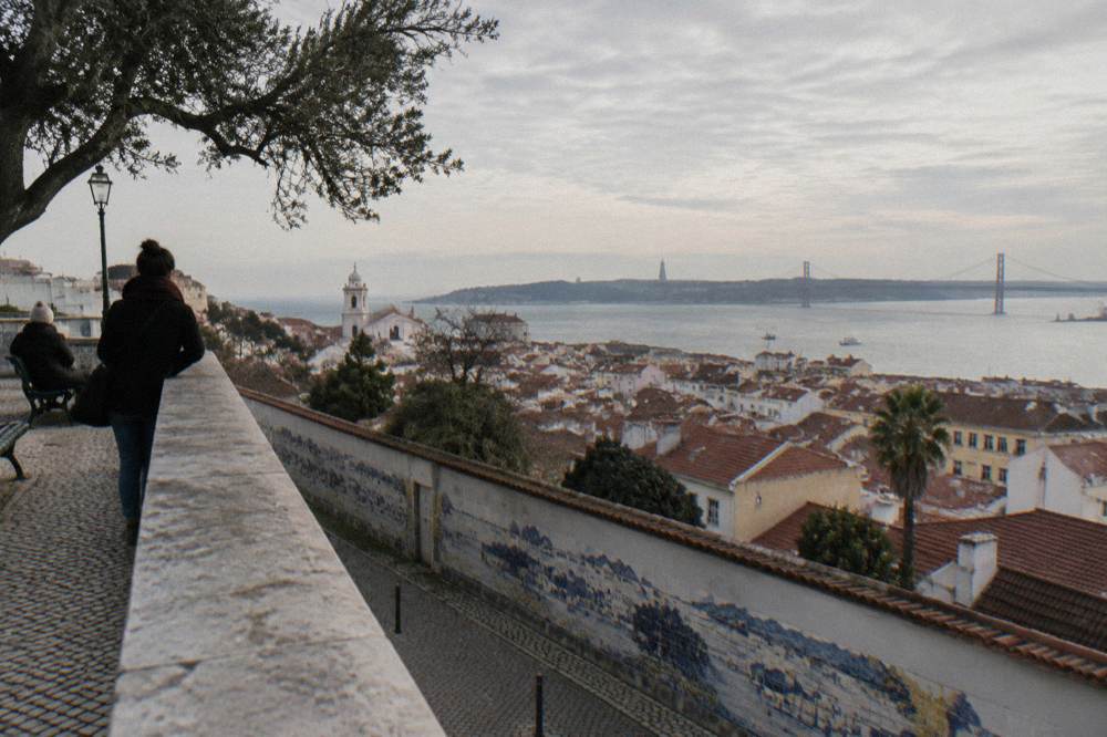 A terrace overlooks tiled Lisbon rooftops, an azulejo wall, the river, and the 25 de Abril Bridge under a cloudy sky.