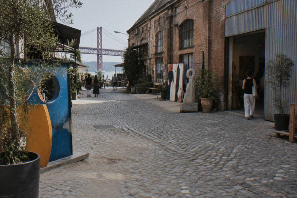 Cobblestoned industrial courtyard with brick and corrugated metal buildings, art installations, a few people, and a red bridge beyond.