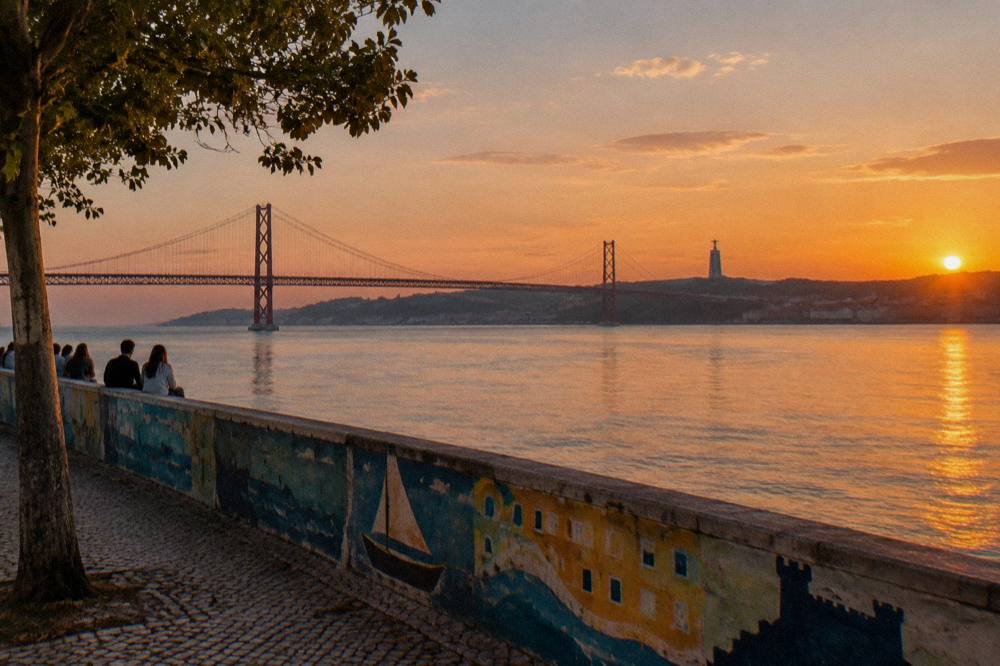 People sit beside a painted riverside wall with the 25 de Abril Bridge and sunset over the Tagus.