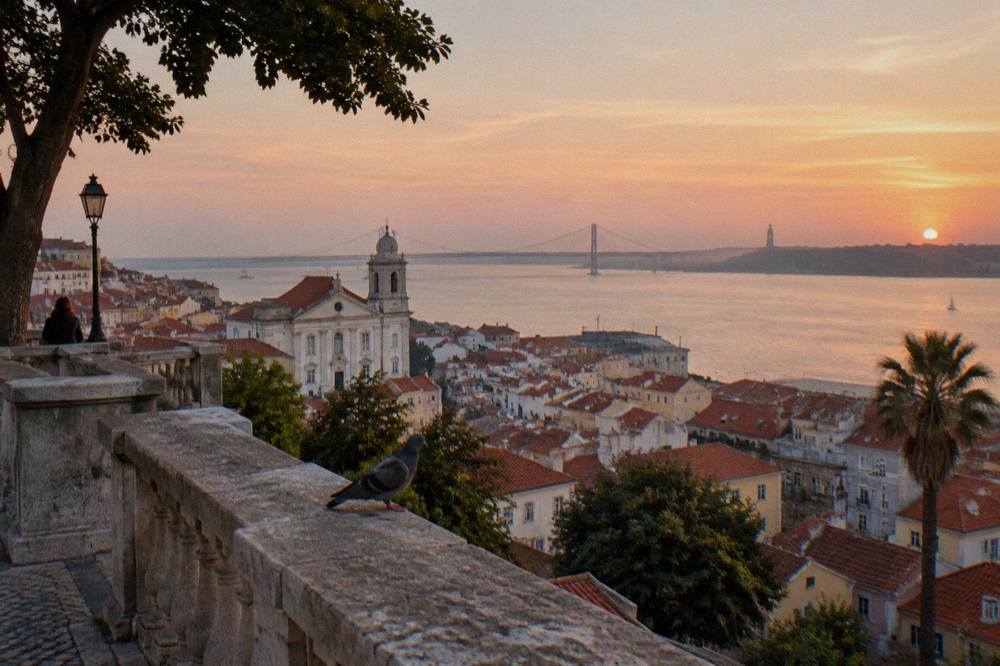 Stone terrace with a pigeon overlooks Lisbon rooftops, the Tagus, a bridge, and a warm sunset sky.