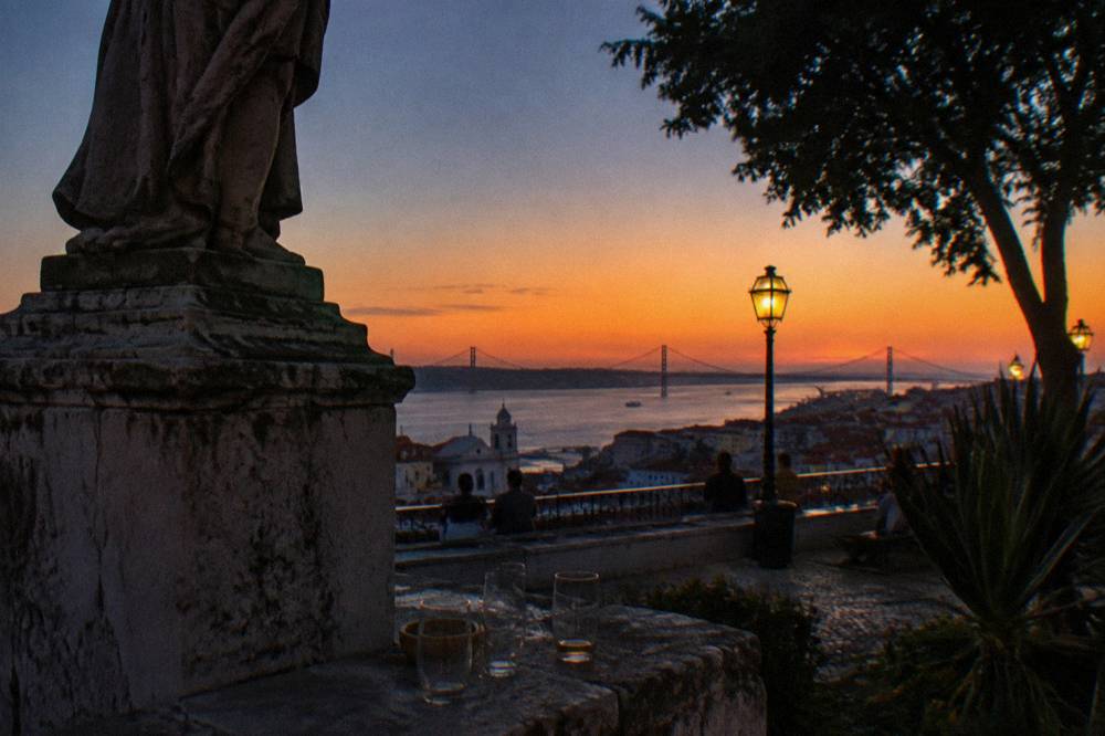 A stone statue pedestal overlooks a dusk river view with a lit lamp, bridge silhouette, and a few glasses on a terrace ledge.