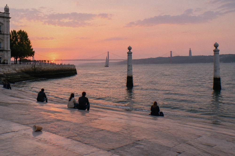 People sit on wide stone steps beside the river at sunset with a sailboat and distant bridge in view.