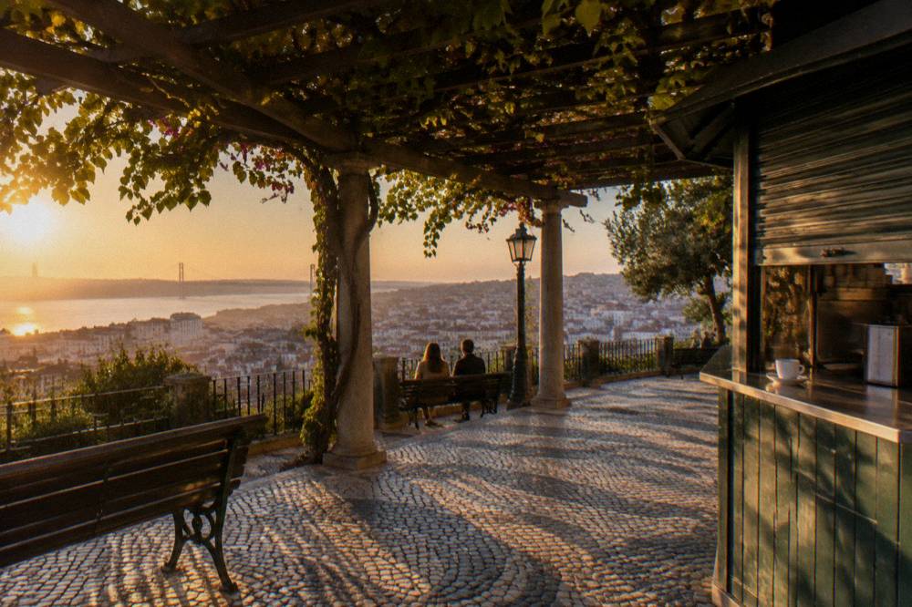 A pergola shaded terrace overlooks Lisbon and the Tagus at sunset, with benches, a kiosk counter, and two people sitting below.