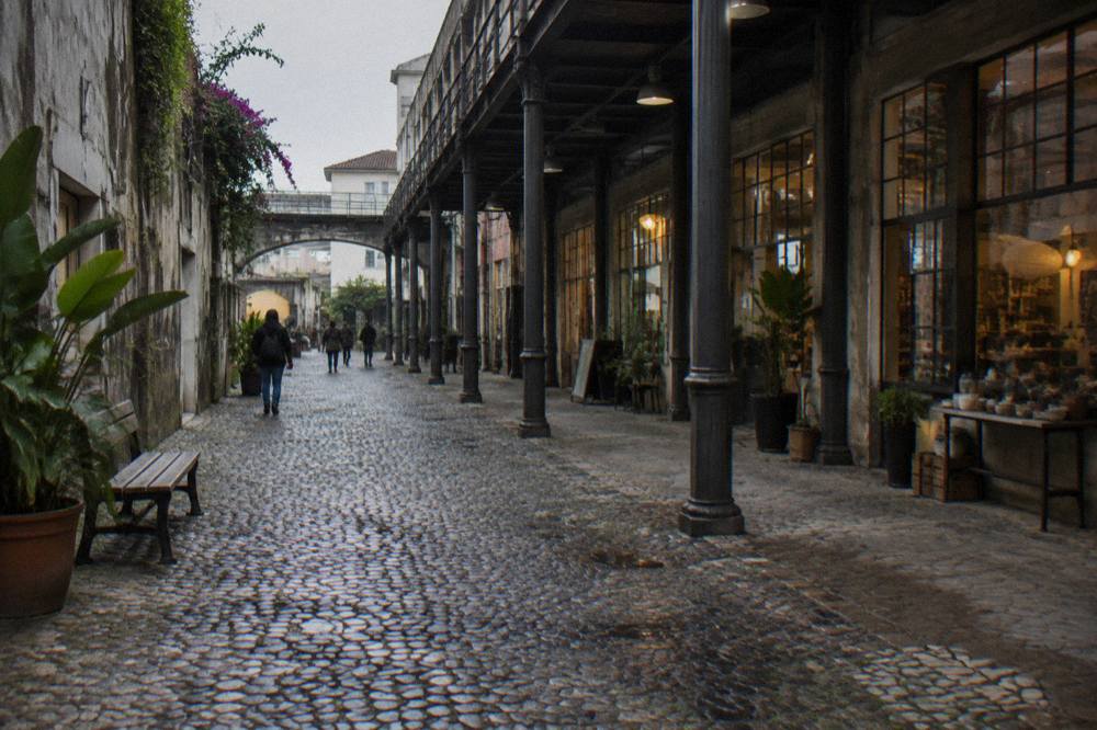 A cobbled lane runs beside cast-iron columns and glass shopfronts under a converted industrial building with a few people walking.