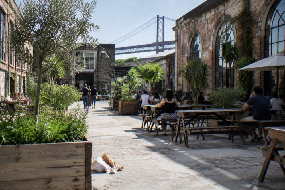 Open courtyard with wooden tables, planters, people seated and walking, and a distant bridge framed by brick factory buildings.