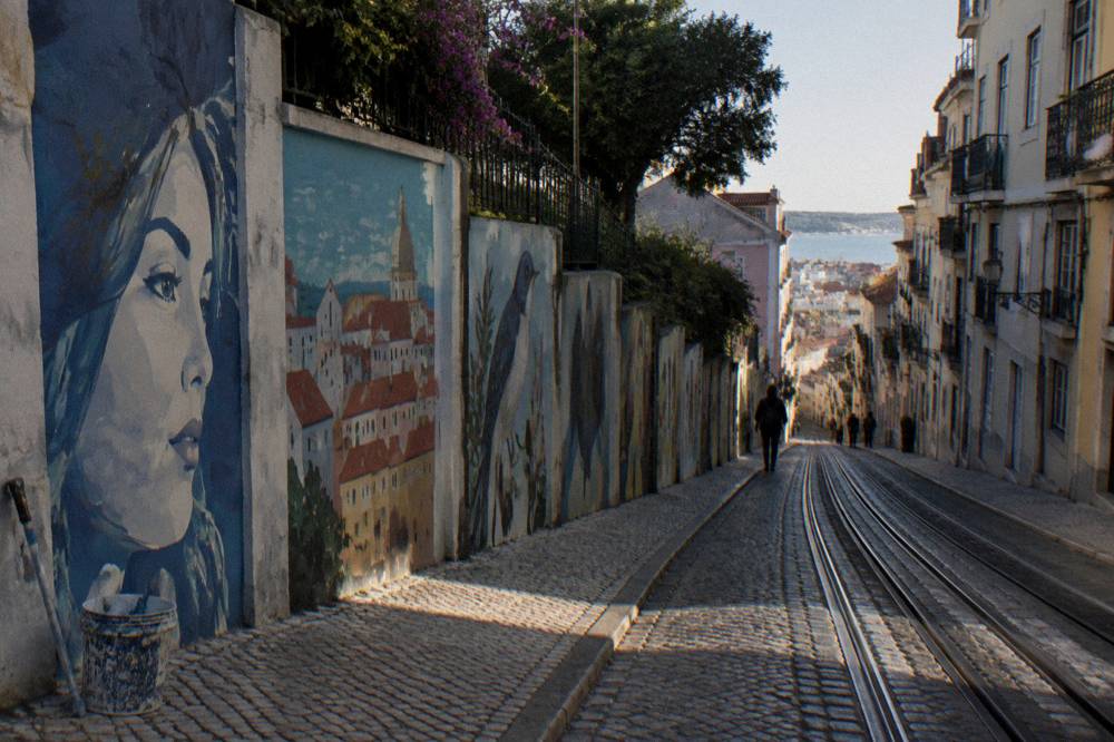 Steep Lisbon street with mural panels, tram tracks, cobbled pavement, a paint bucket, and a distant view over the city to the river.