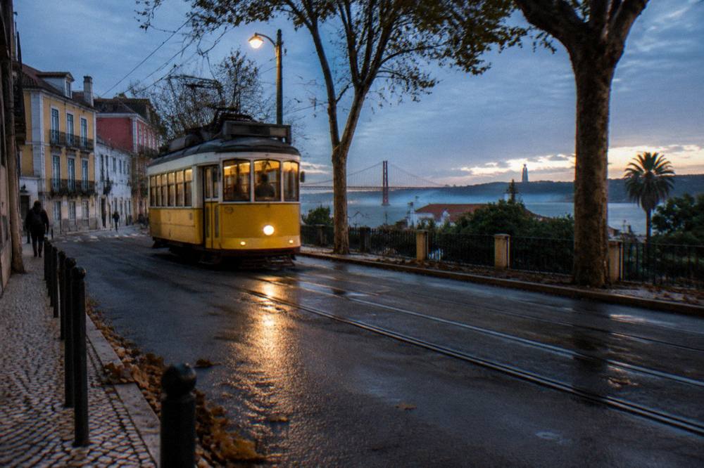 A yellow tram travels along wet street tracks beside trees and riverside buildings at dusk, with the Tagus and a bridge in the distance.