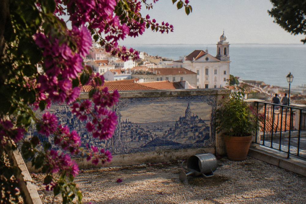 Bougainvillea frames a blue azulejo tile panel above a terrace overlooking Lisbon rooftops and the Tagus.