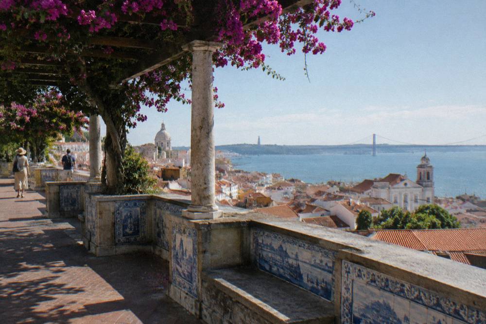 Shaded tiled terrace with bougainvillea overlooks Alfama rooftops, the Tagus River and the 25 de Abril Bridge.