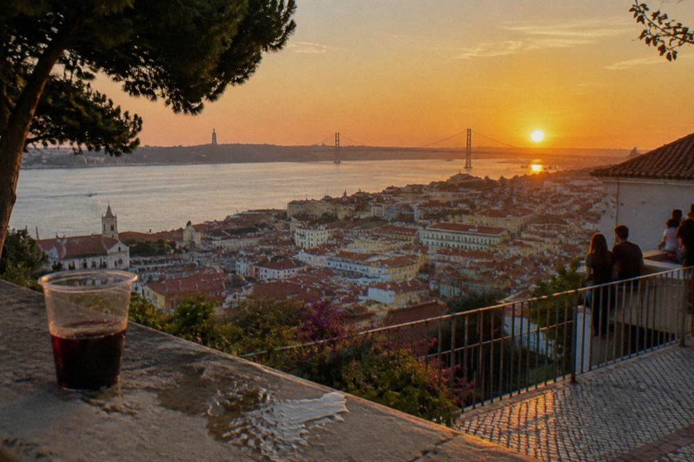 Sunset over Lisbon with the 25 de Abril Bridge, city rooftops, a stone terrace, and a plastic cup of dark drink in front.