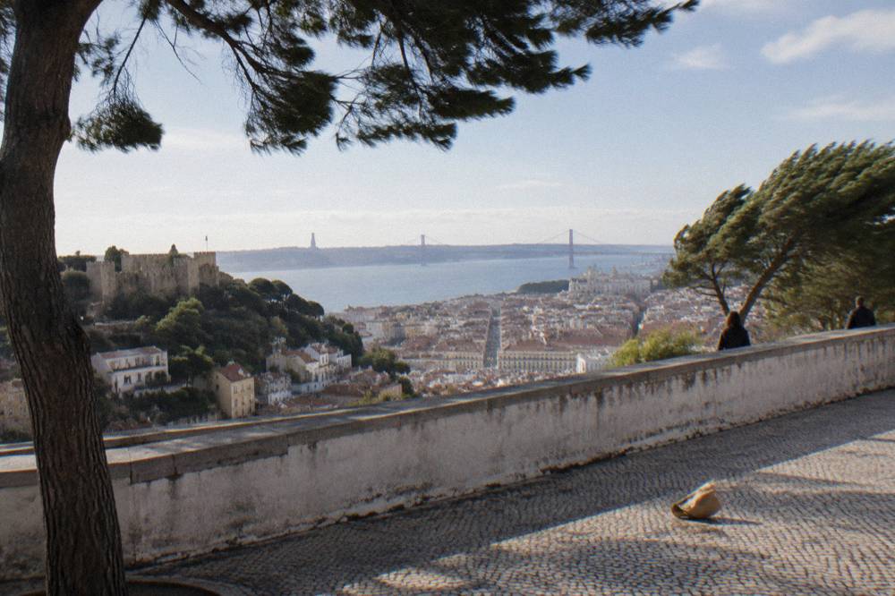 A hilltop terrace with cobbled paving overlooks Lisbon, the castle, the Baixa grid, and the Tagus with a bridge in the distance.