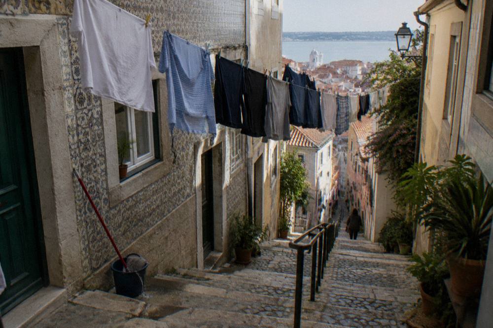 Steep cobbled stair lane between tiled facades with laundry lines overhead, potted plants, and a view toward the river.