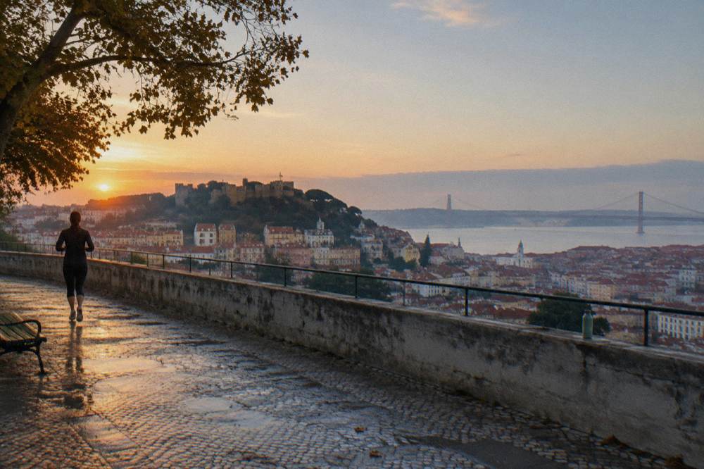 A dawn view from a stone terrace over Lisbon’s castle hill, rooftops, the Tagus and a distant bridge, with a jogger and wet paving.