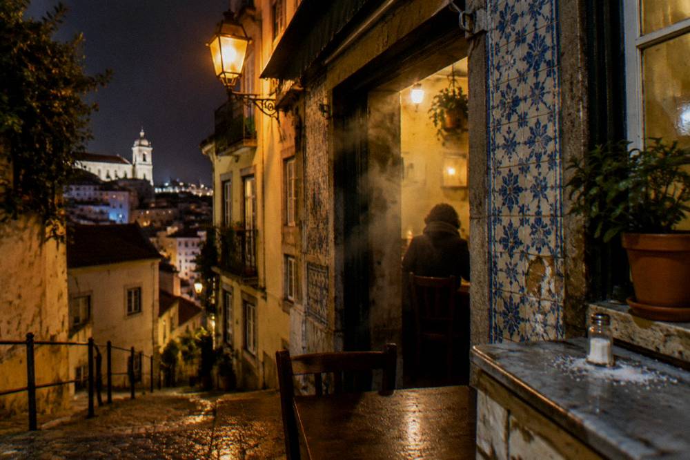 Night view of a tiled Lisbon street corner with a lit eatery, seated figure inside, and a steep cobbled lane below.