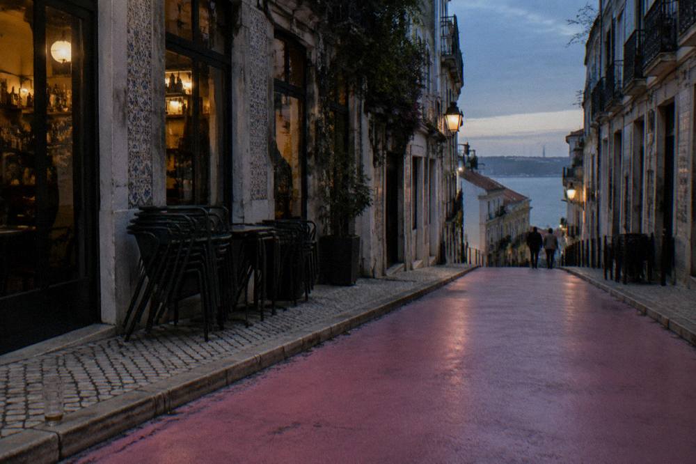A pink street slopes between barfront buildings toward the river at dusk with stacked chairs and a few distant pedestrians.