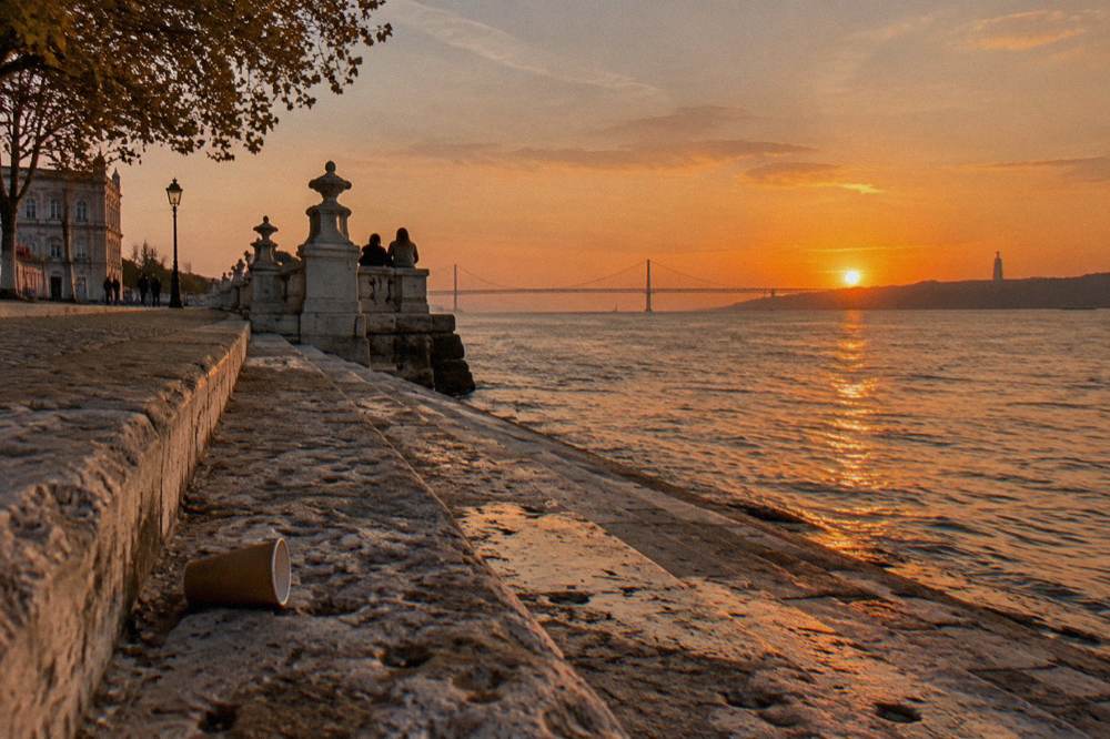 Stone steps beside the Tagus at sunset with orange reflections, a dropped cup, distant people, and a bridge on the horizon.