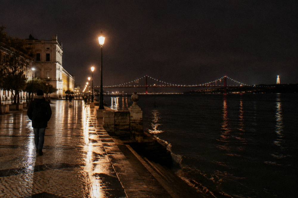 A wet riverside promenade at night with streetlamps, a lone person walking, and a lit suspension bridge across the water.