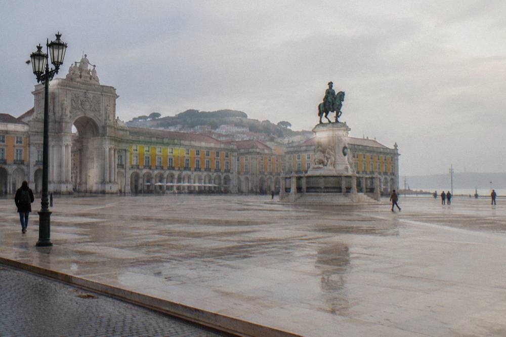 Wide wet plaza with yellow arcaded buildings, a bronze equestrian statue, a tall arch, and a hazy sky by the river.