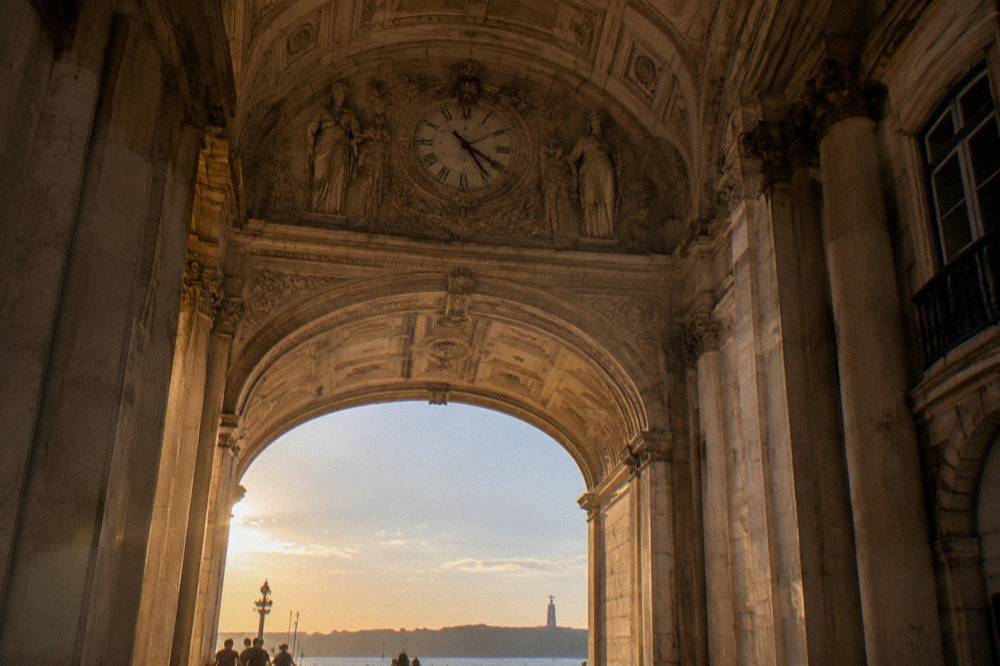 Inside a stone triumphal arch with a clock overhead, looking out toward the Tagus at sunset with small figures below.