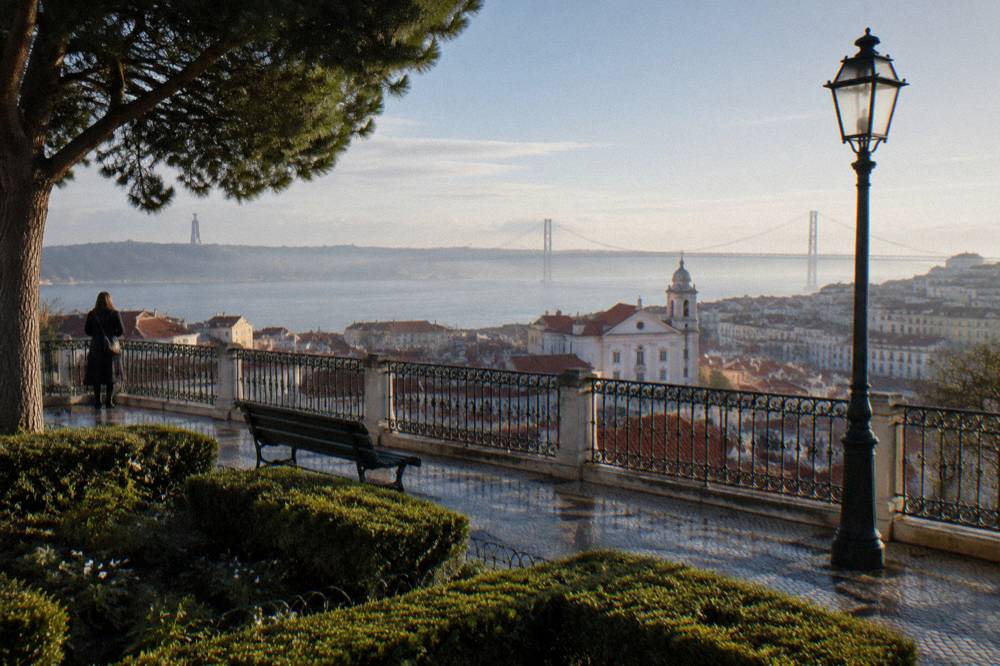 A hilltop terrace with iron railing, benches, hedges, a lamppost, rooftops, and a river and bridge in the distance.