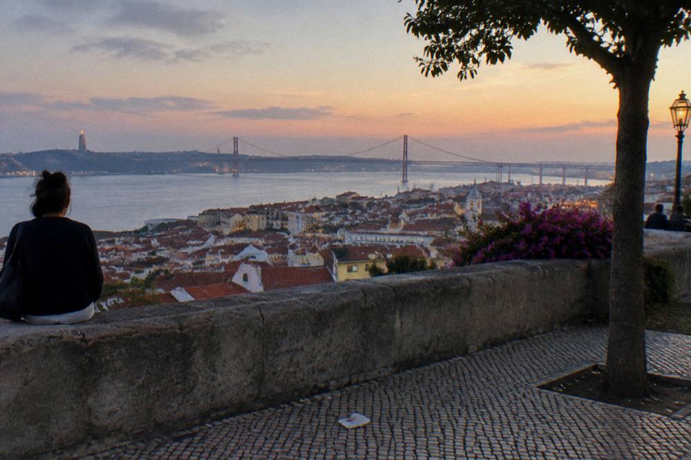 A person sits on a stone terrace above Lisbon rooftops, looking toward the 25 de Abril Bridge over the river at dusk.