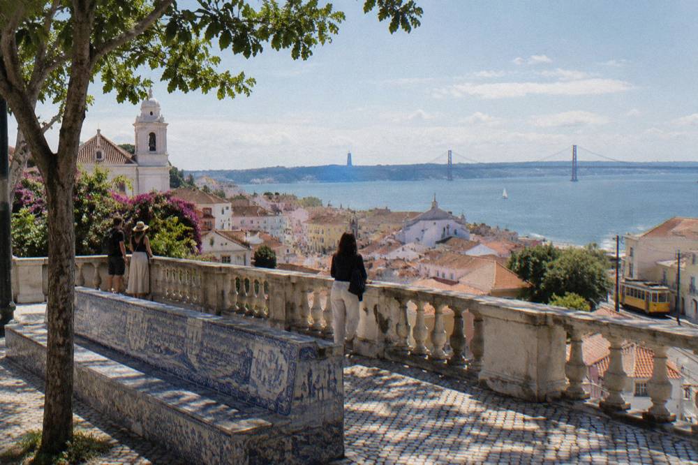 Visitors stand by an azulejo bench at a Lisbon viewpoint overlooking Alfama rooftops, the Tagus River and a yellow tram.