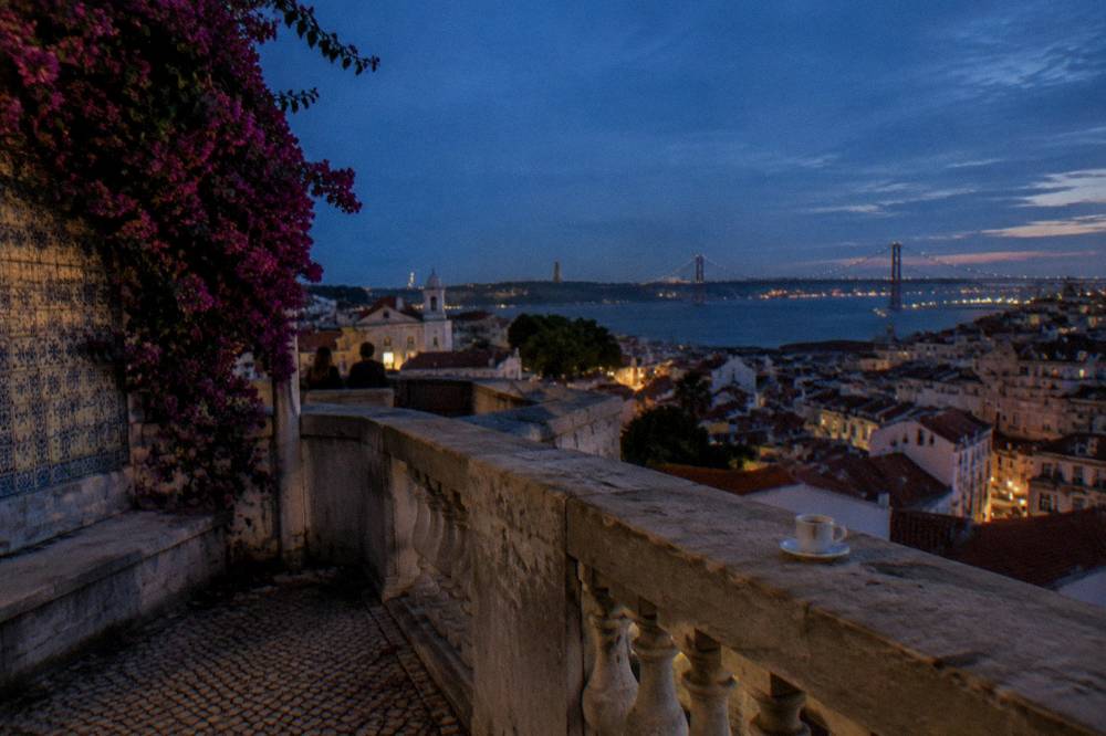 A stone terrace with tiled wall, bougainvillea, a coffee cup, and a dusk view over Lisbon to the river and bridge.