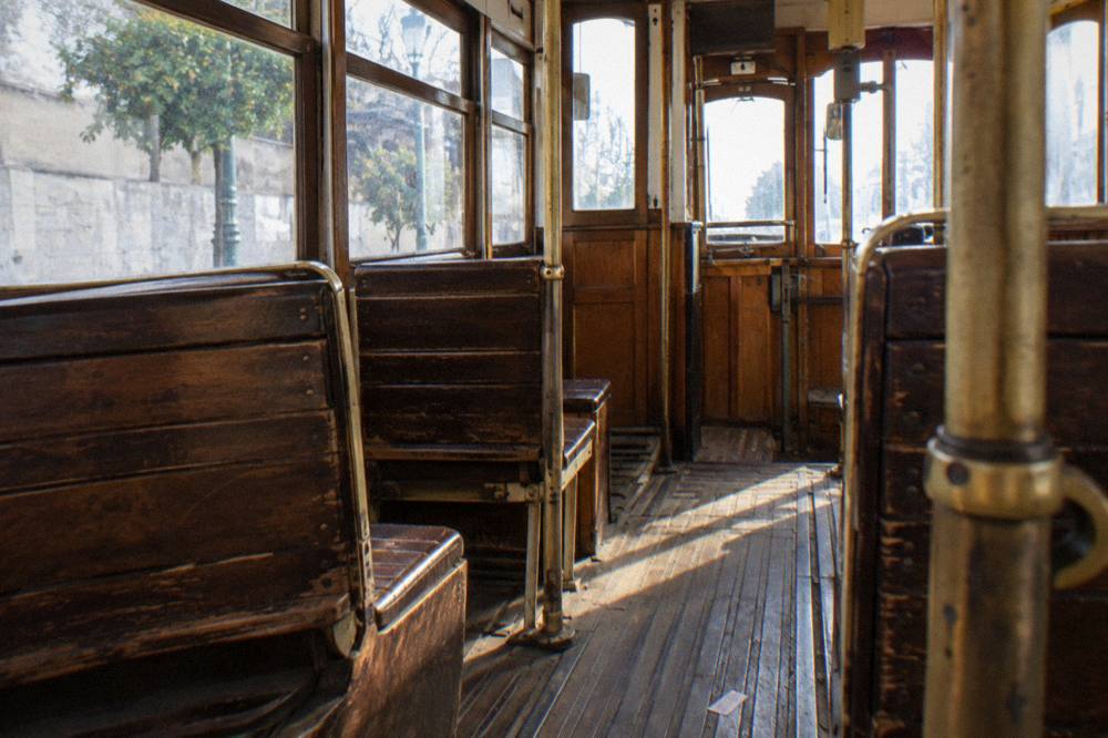 Worn wooden benches and brass poles line an empty vintage tram interior with sunlight across the floorboards.